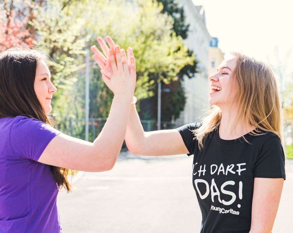 Zwei junge Frauen klatschen sich in der Sonne ab. Sie zeigen Freude und Glück in einem urbanen Umfeld.