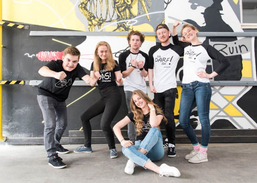 Sechs junge Menschen posieren in T-Shirts mit Aufdrucken, vor einer bunten Graffiti-Wand. Sie zeigen verschiedene Ausdrücke und Gesten.
