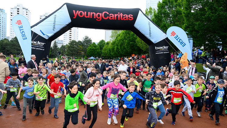 Eine große Gruppe von Kindern läuft unter einem Torbogen beim Jugendlauf von youngCaritas in einem schönen Park.
