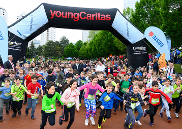 Eine große Gruppe von Kindern läuft unter einem Torbogen beim Jugendlauf von youngCaritas in einem schönen Park.