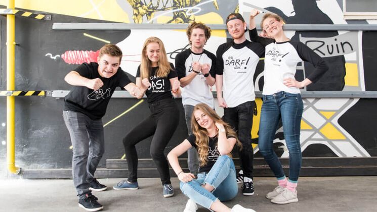 Sechs junge Menschen posieren in T-Shirts mit Aufdrucken, vor einer bunten Graffiti-Wand. Sie zeigen verschiedene Ausdrücke und Gesten.
