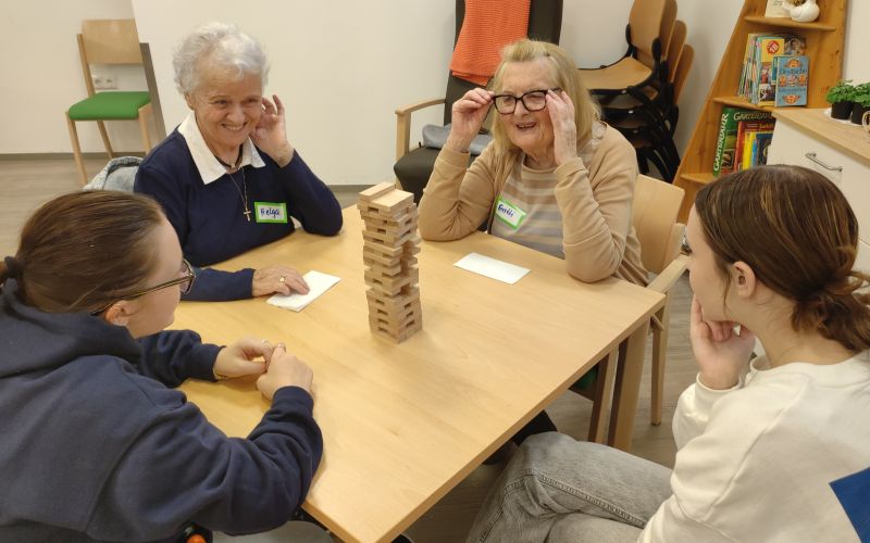 Vier Personen, darunter zwei Seniorinnen, spielen ein Geschicklichkeitsspiel mit einem Holzstapel auf einem Tisch.