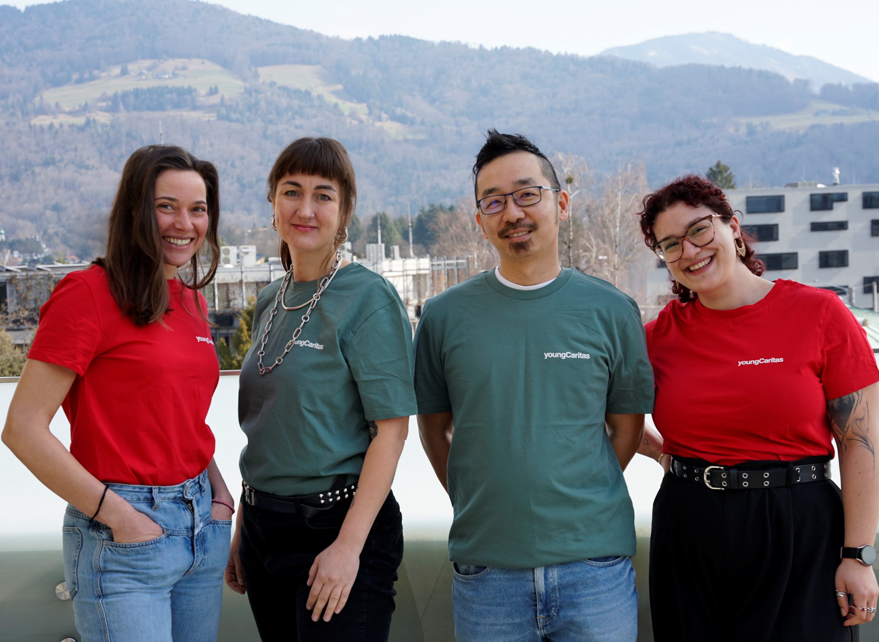 Eine Gruppe von vier Personen in T-Shirts mit dem Logo von Junge Caritas posiert vor einer bergigen Kulisse.