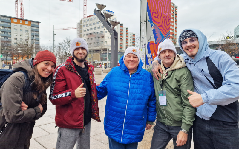Eine Gruppe von fünf Personen posiert freundlich, eingekleidet in Winterjacken und Mützen, in einer städtischen Umgebung.