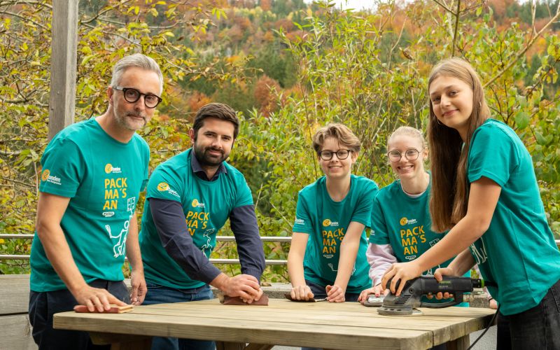 Gruppe von Freiwilligen in grünen T-Shirts arbeitet an einem Holzprojekt in der Natur, um nachhaltig zu gestalten.