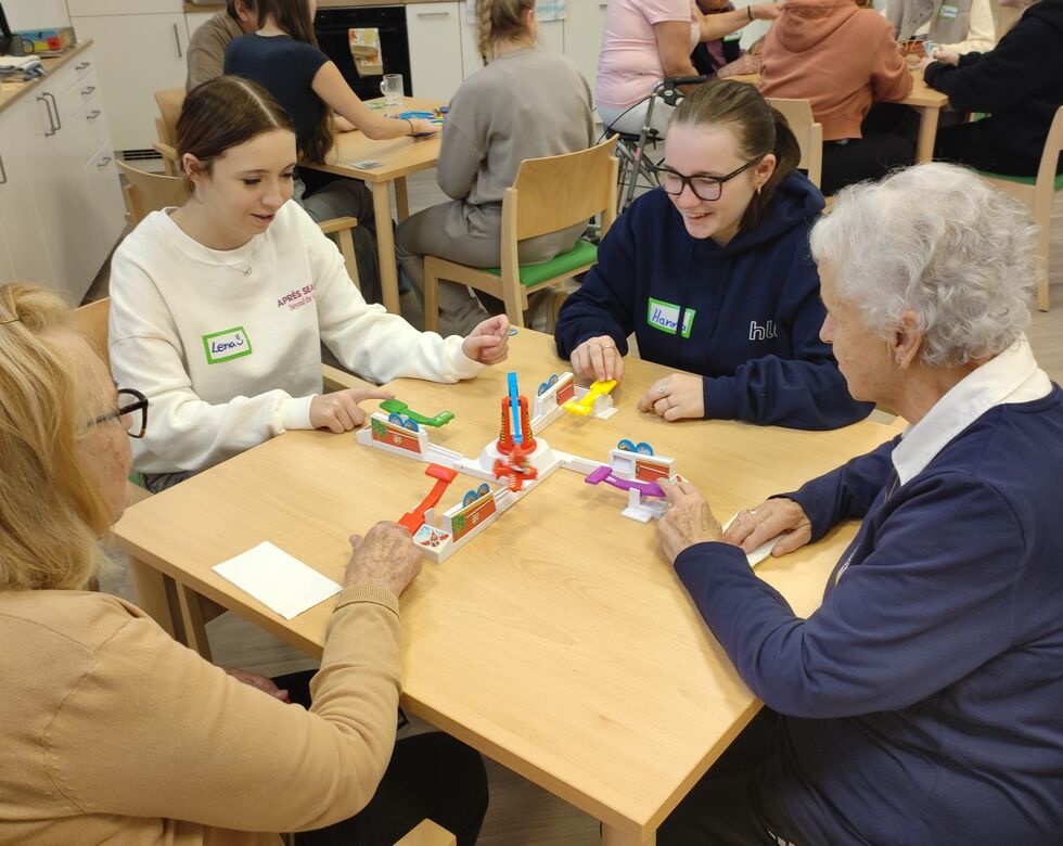 Junge und ältere Menschen spielen gemeinsam an einem Tisch mit einem Spielzeug in einem freundlichen, sozialen Umfeld.