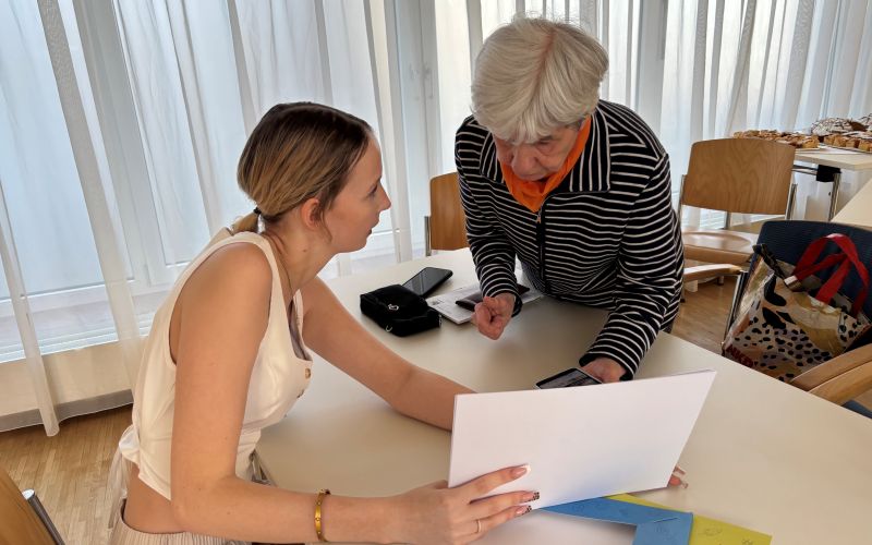 Eine junge Frau zeigt einem älteren Gegenüber Papiere während eines Gesprächs am Tisch in einem hellen Raum.