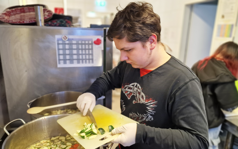 Ein junge Mann schneidet Gemüse in einer Großküche, während andere im Hintergrund mit der Zubereitung von Speisen beschäftigt sind.