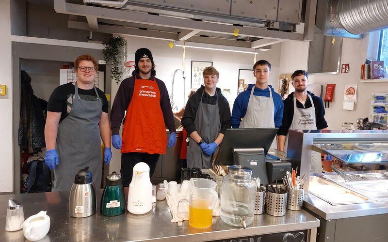 Fünf junge Männer in Kochschürzen stehen in einer Küche. Es gibt Kaffeemaschinen und andere Kochutensilien auf der Theke.