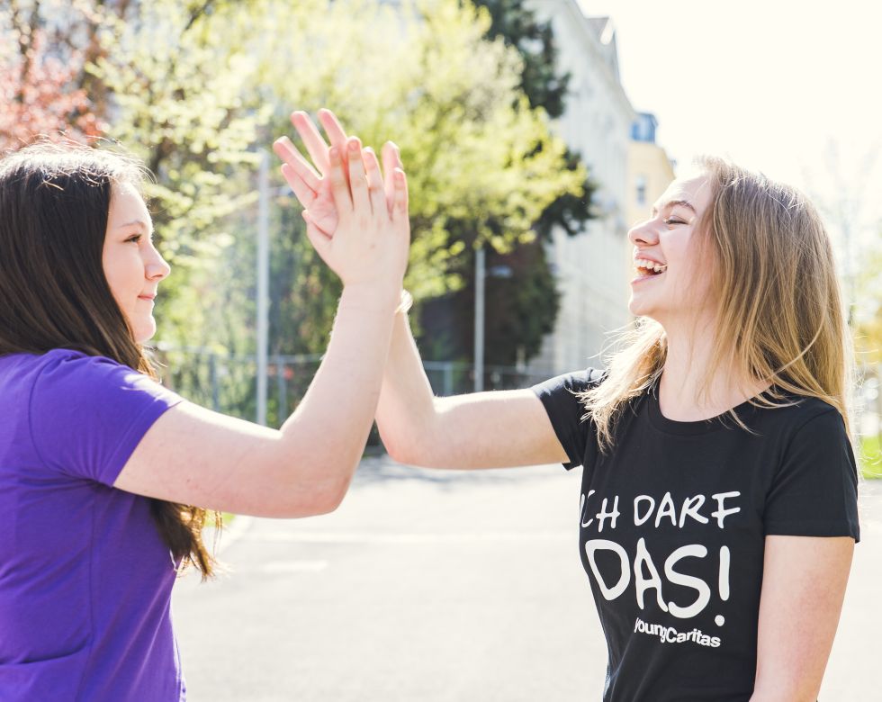 Zwei junge Frauen klatschen sich in der Sonne ab. Sie zeigen Freude und Glück in einem urbanen Umfeld.