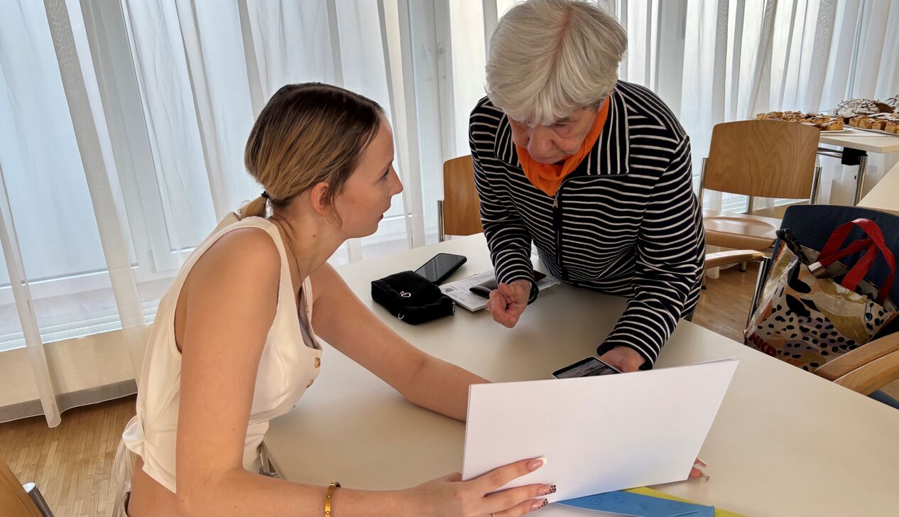 Eine junge Frau zeigt einem älteren Gegenüber Papiere während eines Gesprächs am Tisch in einem hellen Raum.