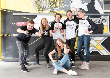Sechs junge Menschen posieren in T-Shirts mit Aufdrucken, vor einer bunten Graffiti-Wand. Sie zeigen verschiedene Ausdrücke und Gesten.