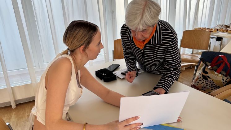 Eine junge Frau zeigt einem älteren Gegenüber Papiere während eines Gesprächs am Tisch in einem hellen Raum.