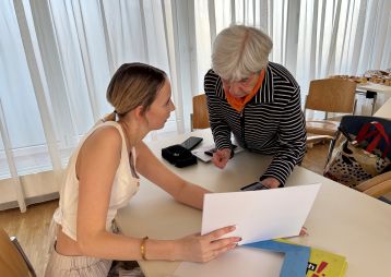 Eine junge Frau zeigt einem älteren Gegenüber Papiere während eines Gesprächs am Tisch in einem hellen Raum.