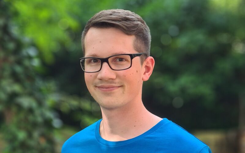 Ein junger Mann trägt ein blaues T-Shirt mit der Aufschrift youngCaritas und lächelt in eine grüne Umgebung.