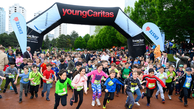 Eine große Gruppe von Kindern läuft unter einem Torbogen beim Jugendlauf von youngCaritas in einem schönen Park.