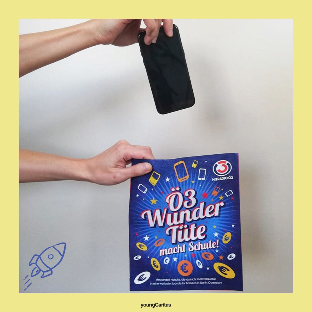 Eine Hand hält ein Smartphone über einem Poster mit dem Text „Ö3 Wunder Tüte macht Schule“. Es geht um Handy-Spenden für Kinder.