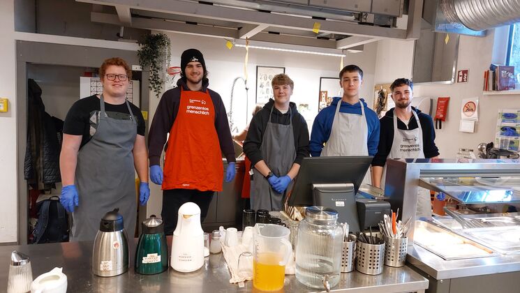 Fünf junge Männer in Kochschürzen stehen in einer Küche. Es gibt Kaffeemaschinen und andere Kochutensilien auf der Theke.