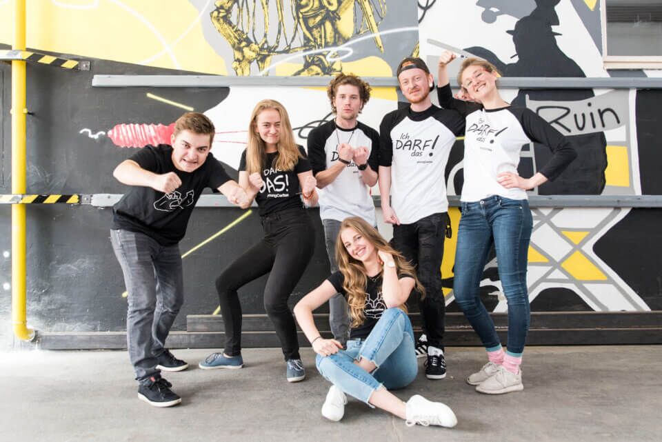 Sechs junge Menschen posieren in T-Shirts mit Aufdrucken, vor einer bunten Graffiti-Wand. Sie zeigen verschiedene Ausdrücke und Gesten.