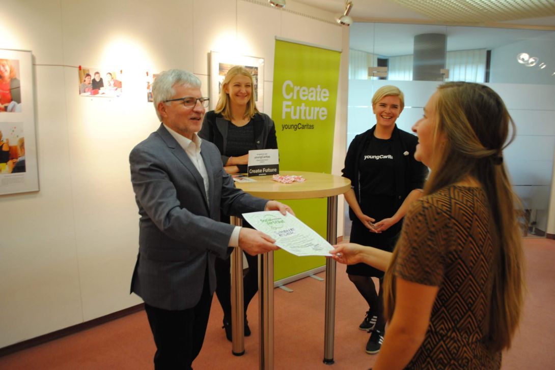 Ein Mann überreicht eine Urkunde an eine Frau, während zwei weitere Personen an einem Tisch stehen.